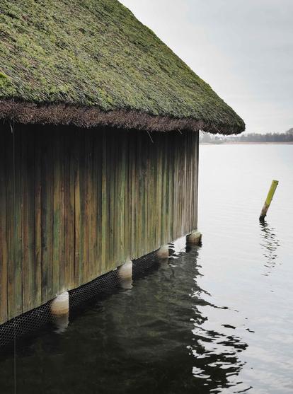 Thatched-roofed wooden building extends over a body of calm water. The roof is covered with moss and the wooden siding shows signs of weathering. Several round wooden supports are partially submerged in the water. The sky is overcast.