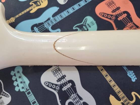 Picture of the back of a guitar neck showing a curved crack running around the neck near the head. The guitar is finished in an ivory white color. It is resting on a black desk mat with various sized graphics of fender jazz basses in white, yellow, blue, green, and orange.