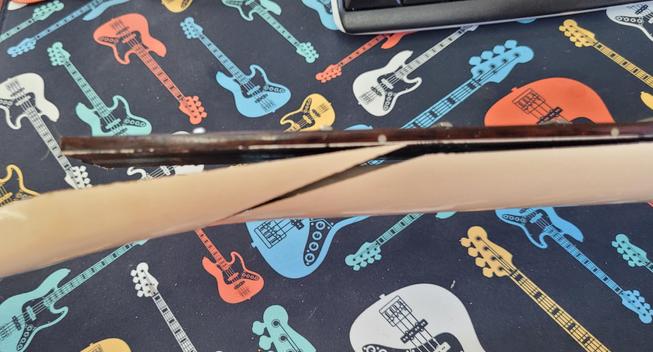 Side view of broken guitar neck. Slanted crack going through the the entire neck and lifting the end of the rosewood fingerboard several millimeters. 
Again, it is resting on the black bass desk mat.