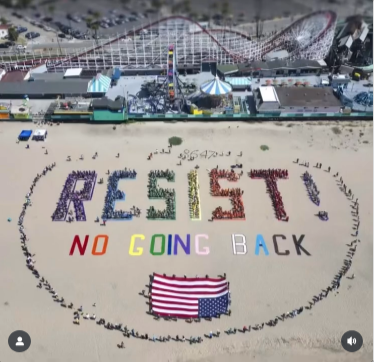 1000 people on the beach today in Santa Cruz. There are people wearing each color of the rainbow standing in lettes that say "RESIST!" in Pride colors. Beneath thta is written "NO GOING BACK." Beneath that, more people are holding a giant upside down US flag of distress. Then there are a whole bunch more people standing in a circle around the whole thing.