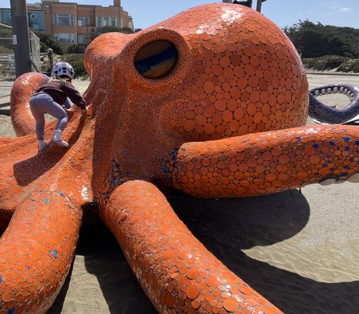 A small child, wearing a helmet and back to the camera, climbs on the octopus sculpture covered in round orange tiles of various sizes. Towards the rear you can see a tentacle with purple tiles rather than orange ones.
