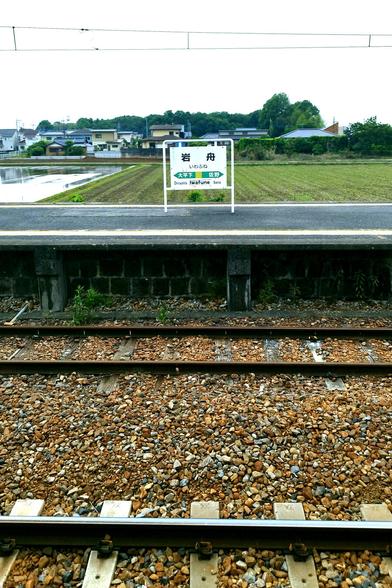 The station has three tracks and two barebone platforms without roofs. This picture was taken from the platform immediately behind the tiny station building looking out over the tracks and the other platform and beyond. Beyond are fields, a few residential buildings, and lots of green. Centered in the shot on the platform is an old station name sign, about chest height. It's not the old, old sign which was in Japanese characters only. It's a bit later when romanization was added and the next stations in either direction.