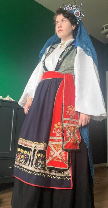 White woman dressed in a white shirt, green bodice, black skirt, blue embroidered apron and red embroidered belt, and a blue crown with a blue veil draping down the back. It is a Swedish folk costume.