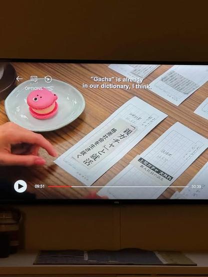 Picture of a TV screen featuring a still with slips of paper with Japanese writing on them, and the closed-caption "'Gacha' is already in our dictionary, I think."