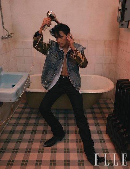 V of BTS wearing his denim jacket with camouflage sleeves with a white shirt underneath, unbuttoned all the way to show his chest and belly button. He is also wearing black pants and cowboy boots. He is sitting on the edge of a white enamel tub looking sideways at the camera while drying his Elvis-styled hair with a blowdryer.