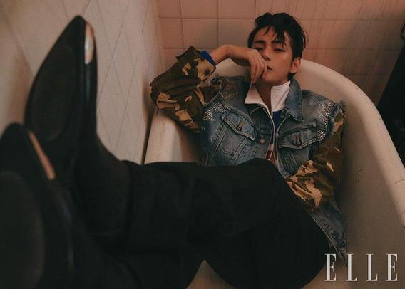 V of BTS lying in a white enamel bathtub, wearing his denim jacket with camouflage sleeves, black pants and cowboy boots. His feet are put up next to the camera. His right elbow is on the edge of the tub, one hand to his lips. He is looking at the camera with heavily lidded eyes.