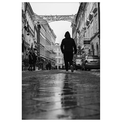A black and white photo of a person in a hooded jacket walking down a wet street in Old Town, Bratislava. The image is taken from a low angle, emphasizing the reflections on the pavement. European buildings line both sides, and festive decorations hang overhead.