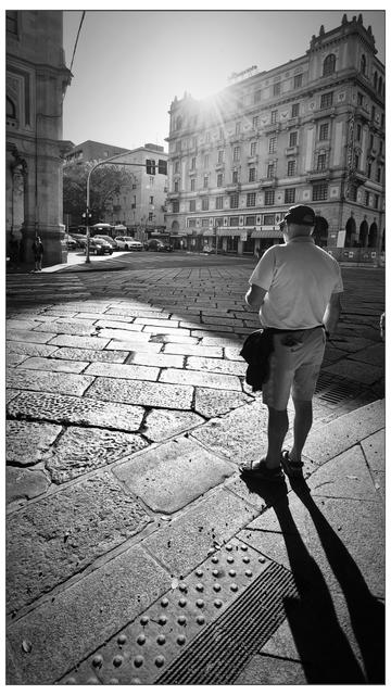 Un uomo in pantaloncini e maglietta, di spalle, attende sul bordo di un marciapiede assolato. La scena è in bianco e nero. Il selciato è ampio e irregolare. In fondo, un palazzo storico è illuminato da un raggio di sole che filtra dall’alto a destra.