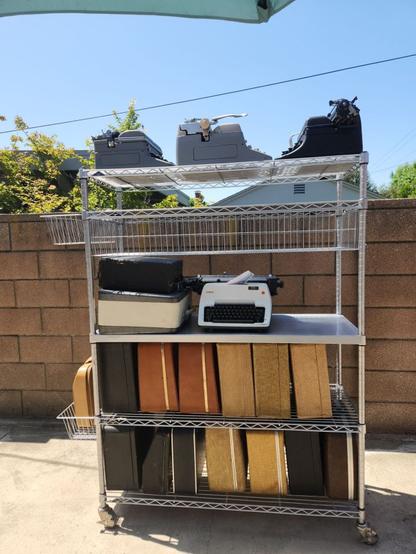 A wire shelving unit on wheels with a variety of 20 typewriters on it. There are four standards and a variety of other portables in a panoply of cases. It sits in a driveway in front of a cinderblock wall.