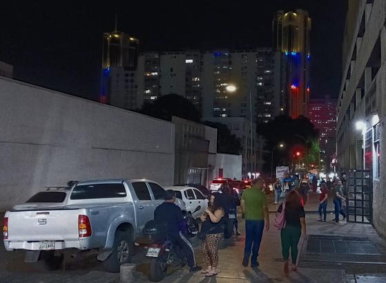 A photo of people going about at night in front of a large residential building. There is a pick-up truck on the left parked on the street and in the background two skyscrapers lit with the colors of the Venezuelan flag are prominent.