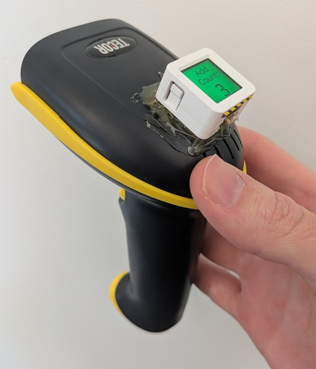 A photo of me holding a yellow and black barcode scanner. There is a white module hotglued to the top of it with a green screen showing "Add Count: 3" in black text.