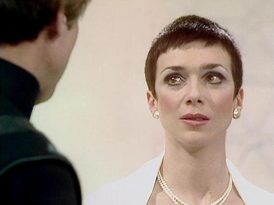 Claude Sonnet 4.0 describes the image as: "I can see this appears to be from a science fiction television series, showing what looks like a dramatic scene between two characters. The image shows a person with very short dark hair wearing what appears to be a white garment and pearl necklace, engaged in conversation with someone whose back is to the camera. The setting appears to be indoors with neutral-colored walls. The lighting and film quality suggest this is from a television production, likely from several decades ago. The scene has a formal, dramatic quality typical of science fiction television of that era."

Blakes7Bot, version 3.9.30.