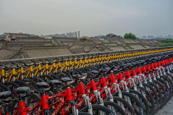A high-angle shot shows numerous rows of parked bicycles in red, black, and yellow. Behind the bicycles is a long, grey brick wall with multiple roofed structures. In the distance, a hazy skyline with various buildings is visible under a grey sky.
