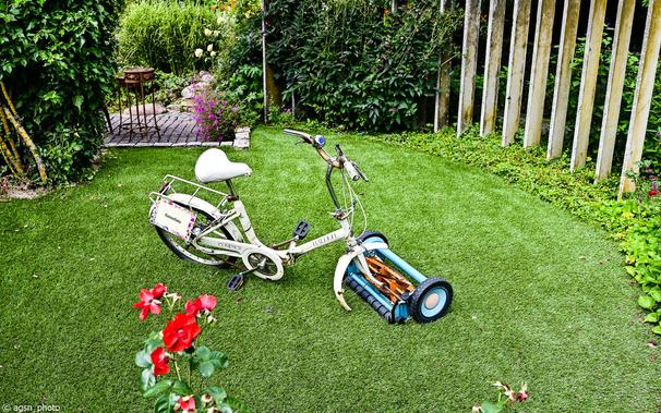 Auf einer Rasenfläche in einem kleinen Garten steht ein altes Fahrrad. Anstelle des Vorderrades wurde ein Handrasenmäher montiert. /
An old bicycle stands on a lawn in a small garden. A push lawnmower has been mounted in place of the front wheel.