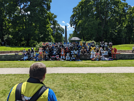 A group of various Fursuiters standing and sitting on a ledge in a park. Lush green trees and an Obelisk centered in the background.