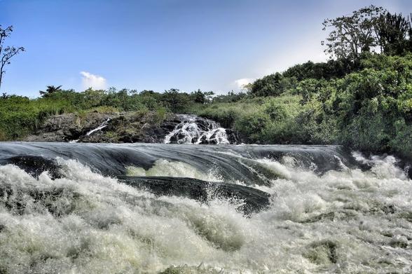 A wide shot of Bujagali Falls in Uganda, showcasing a powerful cascade of white water rushing over dark rocks in the foreground. In the mid-ground, smaller streams of water flow over more rocks, leading up to a lush, green bank of trees and vegetation under a bright blue sky with a few wispy clouds. The sun casts a bright glow on the right side of the image, illuminating the trees and creating a sense of natural grandeur.