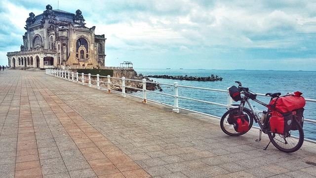 The landscape-orientated image displays the Constanța Casino, a large, ornate building, on the left side, situated next to a wide paved promenade. A white metal railing separates the promenade from the calm, blue sea on the right. The sky above is cloudy, with shades of white and light blue. In the foreground on the right, a touring bicycle with red panniers is parked on the promenade, facing the sea. In the distance, several large ships are visible on the horizon. The overall scene suggests a coastal setting with historical architecture and an active waterfront.
