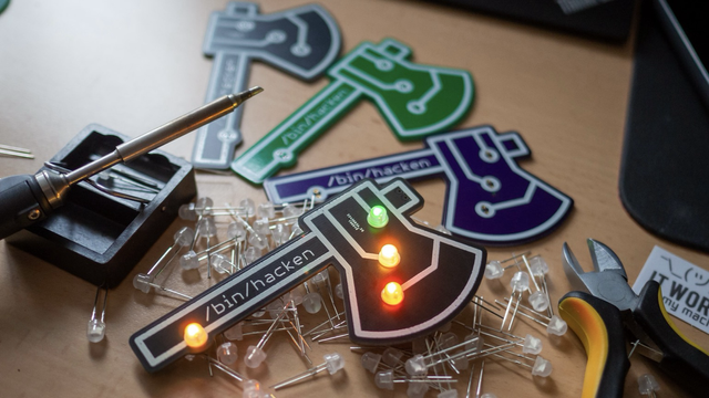 Vier Badges in der Form der /bin/hacken-Axt liegen auf einem Tisch. Eines davon mit leuchtenden LEDs ausgestattet. Die anderen sind noch nicht fertig, mehrere LEDs liegen verteilt, außerdem ein Lötkolben und eine Zange.