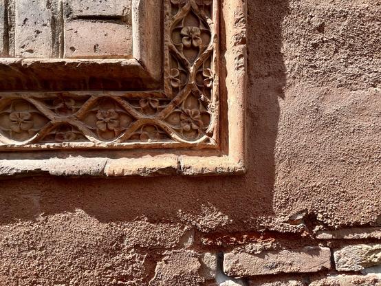 This is a bit difficult to explain, but here we go: detail shot of a wall, with a series of rectangular corners, expanding from the top left hand of the corner of the image, like a stack of paintings or papers or books laid one on top of each other, in order of size. Or, like a chart showing different paper sizes. The first and smallest, in the top left corner, has three small bricks, standing out from the wall, in a sort of frame. Next is an ornate carved relief border, with ribbons undulating to form circles which have flowers inside them; this too is contained within a narrow frame, like that of a painting, which casts a dark and jaggedy-edged shadow . The rest of the wall is uneven textured plaster, which has fallen away in the bottom right hand corner to expose some rough brick. The whole is in shades of pastel red, and basking in sunshine.