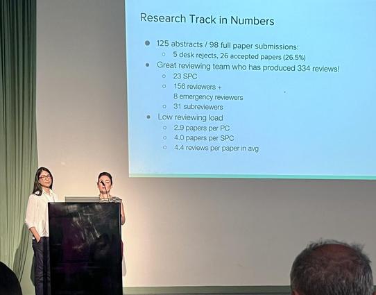The image shows a presentation slide titled "Research Track in Numbers" displayed on a projector screen. The slide details the statistics of a research track, including the number of abstracts and full paper submissions, the acceptance rate, and the reviewing team's performance. It states that there were 125 abstracts and 98 full paper submissions, with 5 desk rejects and 26 accepted papers, representing a 26.5% acceptance rate. The reviewing team consisted of 23 Senior Program Committee (SPC) members, 156 regular reviewers, 8 emergency reviewers, and 31 subreviewers. The reviewing load was low, with an average of 2.9 papers per PC, 4.0 papers per SPC, and 4.4 reviews per paper. The slide is presented by two individuals standing behind a podium, with one person speaking into a microphone. The background is a plain wall with a green curtain on the left side.

Provided by @altbot, generated privately and locally using Ovis2-8B