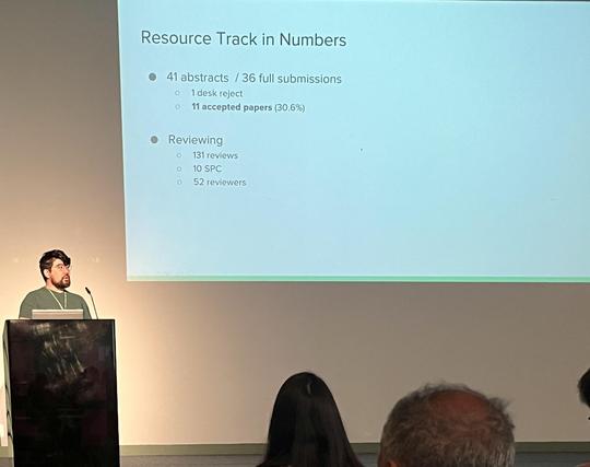 The image shows a presentation slide titled "Resource Track in Numbers" displayed on a large screen. The slide contains bullet points with numerical data. The first bullet point states "41 abstracts / 36 full submissions," with sub-points indicating "1 desk reject" and "11 accepted papers (30.6%)." The second bullet point is labeled "Reviewing" and includes "131 reviews," "10 SPC," and "52 reviewers." A person is standing at a podium to the left of the screen, wearing a green shirt and a lanyard, with a microphone in front of them. The audience is partially visible, with the backs of their heads shown. The room appears to be a conference or seminar setting, with a plain background and a projector screen.