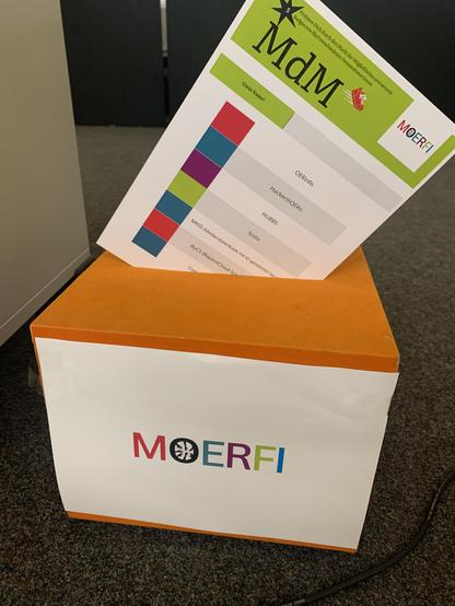 Kiste mit "MOERFI-Aufschrift". Darin steckt ein Blatt, das auf den Markt der Möglichkeiten hinweist.