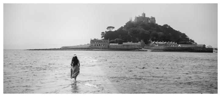A woman with her back to camera walking out into the sea. Just visible under the water is a cobble stone path she is walking along. In the distance a small island is rising from the sea. It has a castle on the top of a hill rising steeply from the water.