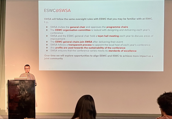 The image shows a presentation slide titled "ESWC[@]SWSA" displayed on a large screen. The slide outlines the oversight rules that SWSA will follow with ESWC, similar to those at ISWC. The text includes bullet points detailing the roles and responsibilities, such as inviting the general chair, approving program chairs, and holding town hall meetings for improvement. It also mentions the transparent process for appointing local hosts, using profits for sustainability, and ensuring the conference series meets standards of excellence. The slide concludes with a note about exploring opportunities to align ESWC and ISWC for greater impact as a joint community.

In the foreground, a person stands at a podium, presumably delivering the presentation. The audience is partially visible, with a few individuals facing the speaker. The setting appears to be a conference room with a plain wall and a projection screen. The overall atmosphere suggests a formal presentation or meeting.

Provided by @altbot, generated privately and locally using Ovis2-8B