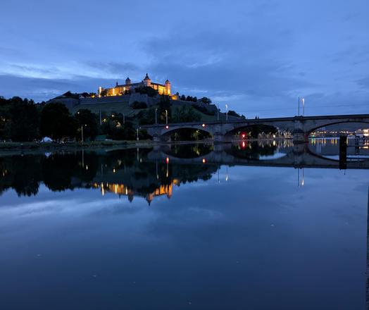 Die beleuchtete Festung Marienburg in Würzburg bei Nacht vom anderen Mainufer aus aufgenommen. Die Festung und der Berg, auf dem sie steht, spiegeln sich im nächtlich blauen, glatten Wasser des Mains