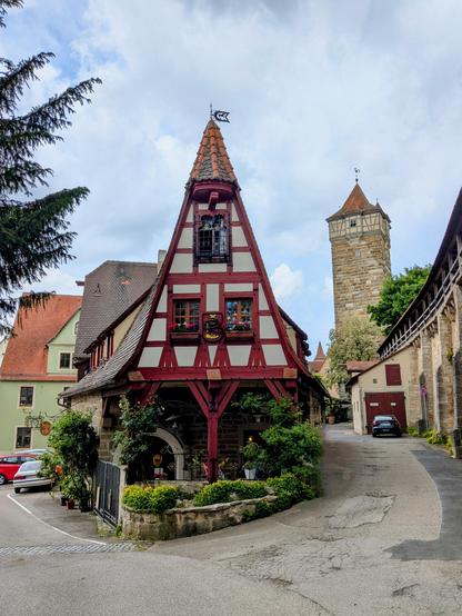 Ein Fachwerkhaus mit roten Balken und sehr spitzem Dach in Rothenburg, danaben verläuft die mittelalterliche Stadtmauer, im Hintergrund sieht man einen Turm in der Stadtmauer