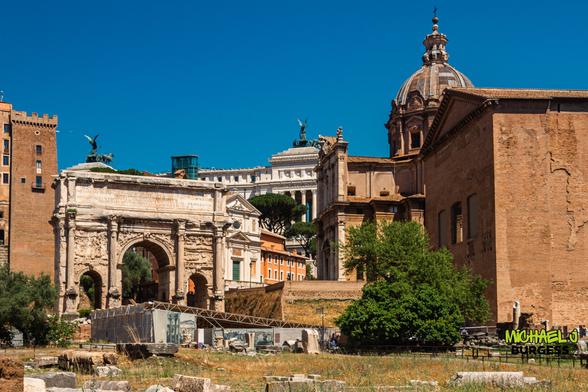A wide-angle view of the Arch of Septimius Severus in Rome's Roman Forum, with classical buildings and a dome rising in the background under a clear blue sky.