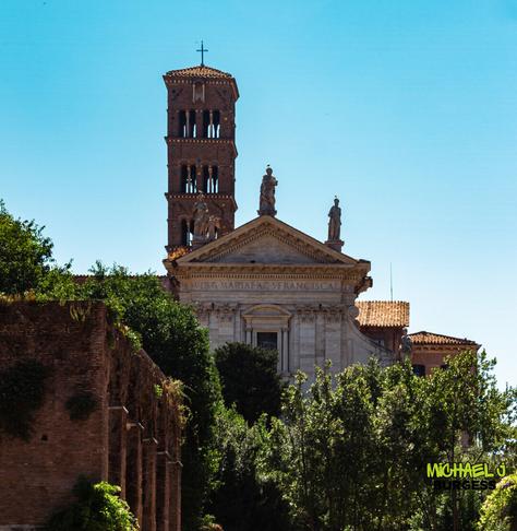 Frontal view of the Basilica of Santa Francesca Romana with its tall Romanesque bell tower and statues atop the pediment, framed by trees and ancient Roman ruins under a clear blue sky.