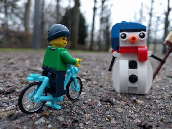 Eine Lego Minifigur mit Mütze und grünem Pulli sitzt auf einem Lego-Fahrrad. Neben ihr steht ein aus Lego gebauter Schneemann. 
Die Szene spielt auf einem geteerten Weg mit sichtbaren Steinchen. Im Hintergrund sind karge Bäume und (mit viel Phantasie) das Karlsruhe Schloss zu sehen.