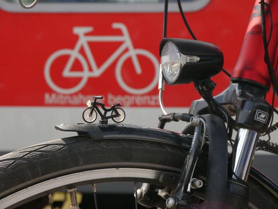Zu sehen ist der obere Teil des Vorderrades eines Fahrrads mit Schutzblech, Bremse, Federgabel und Scheinwerfer. Auf dem Schutzblech ist ein Lego-Fahrrad als Kühlerfigur angebracht. Im Hintergrund ist unscharf ein weißes Fahrrad auf einem roten Streifen gedruckt auf einem Zugwaggon zu sehen.