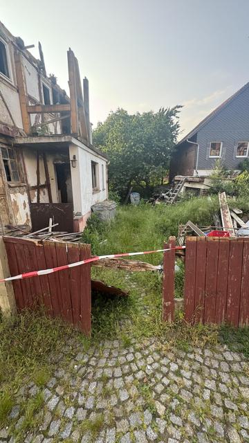Ein verlassenes Grundstück mit einer teilweise eingestürzten Struktur und überwucherter Vegetation. Zerbrochene Holztore und Bauschutt sind im Hof neben Mülleimern zu sehen. Im Hintergrund sind zwei Häuser zu sehen.