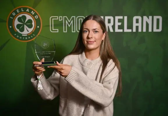 Photograph of a woman in a cream jumper holding a glass trophy aloft. She is stood in front of a green background