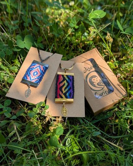 Photo d'un pendentif tissé aux couleurs trans et d'un porte-clés arc-en-ciel de la collection kaléidoscope queer posés dans l'herbe, à coté d'un packaging avec le logo de la marque.