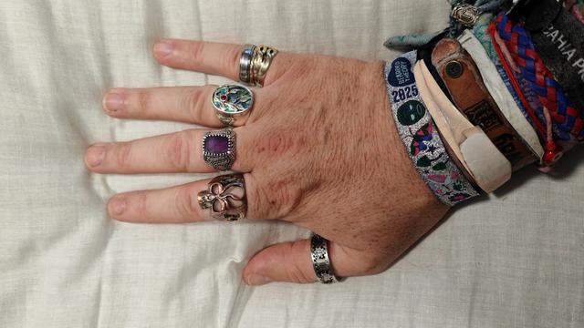 A man's hand displaying five ornate rings across his fingers - including a chunky gold band, a green gemstone signet ring, a purple stone surrounded by intricate metalwork, a copper-toned floral design, and a patterned silver band on his thumb. Multiple colourful fabric wristbands complete the understated look against white bedding.