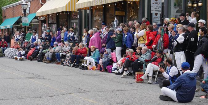 Several dozen people standing and sitting along a street, watching a performance out of frame to the left. Several of them are in morris dancing costume.