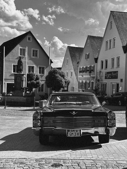 Alt-text (English):
A black-and-white photograph of a vintage Cadillac parked in a European town square. The car faces the camera, prominently displaying its grille and classic design. In the background are traditional, steep-roofed buildings and a statue in front of a small fountain or monument. One of the buildings has a sign reading “Café Franco.” The sky is partly cloudy, creating dramatic lighting and contrast in the image. The scene evokes a nostalgic atmosphere, blending American automotive history with a quaint European setting.
