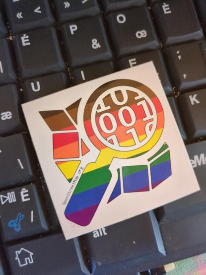 Photo d'un autocollant posé sur un clavier noir, il représente le logo d'openstreetmap, une loupe posée sur une carte, en dégradé arc-en-ciel (8 bandes horizontales, de haut en bas : noir, marron, rouge, orange, jaune, vert, bleu, violet) sur fond blanc