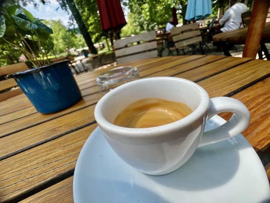 Eine Espresso in einer kleinen, weißen Tasse, auf einem Holztisch in einem Café an einem Park im Hintergrund