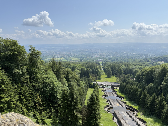 Blick vom Herkules nach Kassel: sehr viel Wald, durchschnitten von einer langen Linie durch die Wasserbecken und deren Sichtachse, die sich bis in die Stadt fortsetzt. Blauer Himmel mit einigen Wölkchen.