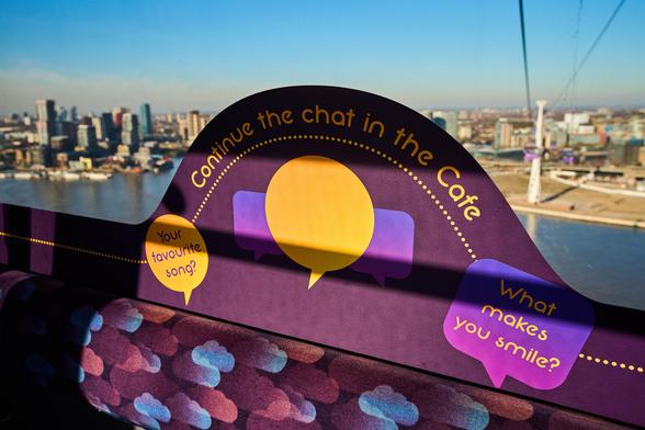 The image shows the interior of a cable car with a sign encouraging conversation. The sign has a curved top and is positioned above the seat. It features three overlapping speech bubbles in purple and yellow. The largest bubble in the centre is yellow, flanked by two smaller purple ones. The words "Continue the chat in the Cafe" are written in a playful, curvy font near the top of the sign. The cityscape visible through the window includes buildings and a river, suggesting an urban setting.

Photo by TfL
