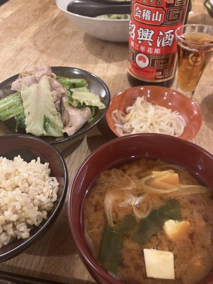 A meal featuring a bowl of miso soup with vegetables, a side of cooked pork and cabbage, steamed bean sprouts, and brown rice. A bottle of drink is visible alongside a small glass. The setting appears to be a wooden table.
