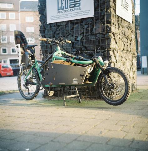 Ein grünes Káro der  Bremer Firma VeloLab vor einen mit Steinen befüllten Gitter im Abendsonnenschein.