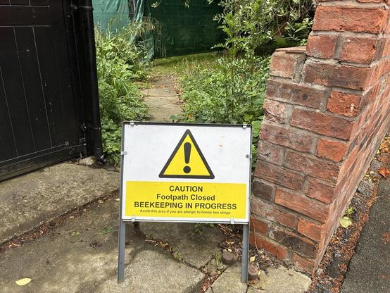 A sign indicating "Caution: Footpath Closed, Beekeeping in Progress" is placed near a gate. The surrounding area is overgrown with grass and plants.