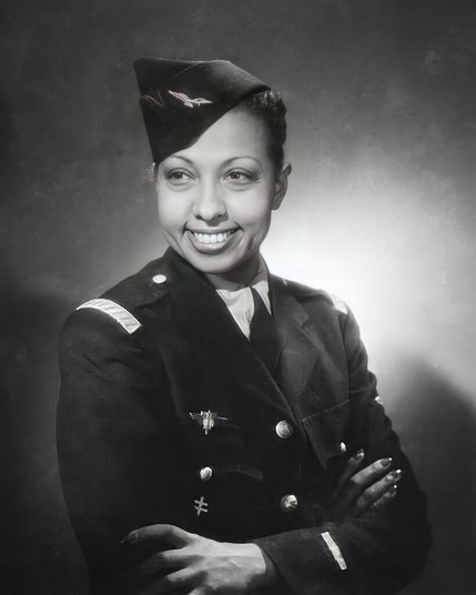 Photo en noir et blanc de Joséphine Baker, prise de profil. Elle est vêtue d’un uniforme militaire sombre, décoré de médailles, avec les bras croisés. L’arrière-plan est sombre et légèrement flou.