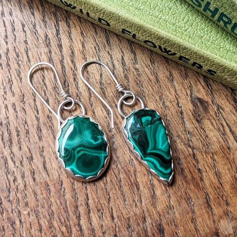 A pair of earrings made of mismatched malachite stones set in silver on ear wires. They are resting on a wooden surface, next to some green hardback books.