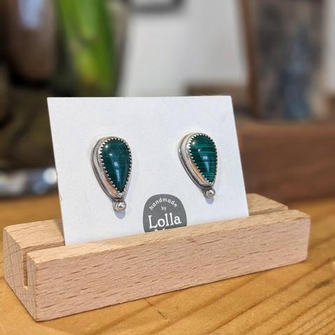 A pair of earrings mounted on a white card bearing the LollaMac logo, in a wooden holder. The earrings are upside-down teardrop shaped malachite, in a silver setting with a small silver ball beneath.